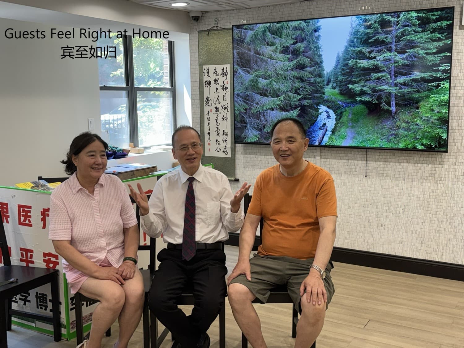 李医生与两位来宾坐谈，背景大屏为森林风景、旁有书法卷轴